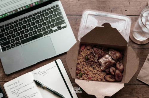 Flat-lay of a takeaway jollof rice lunch on a wooden desk next to an open laptop and notebook