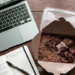 Flat-lay of a takeaway jollof rice lunch on a wooden desk next to an open laptop and notebook