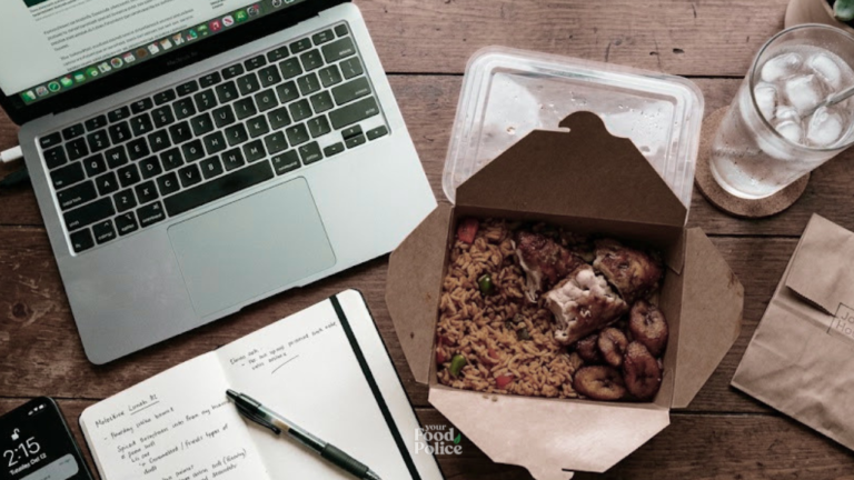 Flat-lay of a takeaway jollof rice lunch on a wooden desk next to an open laptop and notebook
