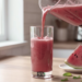Pour of fresh watermelon smoothie from a blender into a tall glass with a side of sliced fruit and lime wedges.