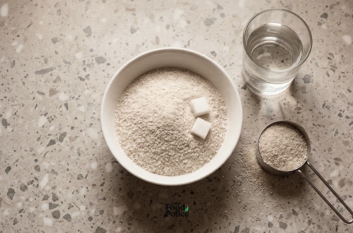 Overhead flat-lay of a white bowl filled with garri granules and sugar cubes, next to a glass of water and a measuring cup.