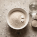 Overhead flat-lay of a white bowl filled with garri granules and sugar cubes, next to a glass of water and a measuring cup.