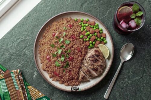 Overhead view of a jollof rice and grilled chicken meal with zobo drink on a dark stone surface next to an colorful ankara napkin.