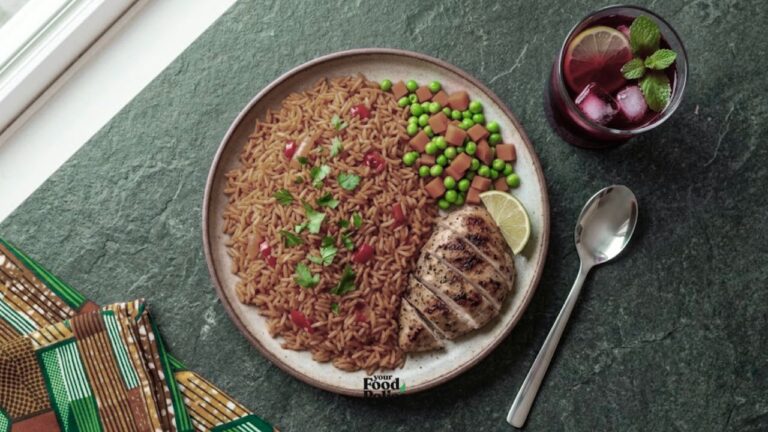 Overhead view of a jollof rice and grilled chicken meal with zobo drink on a dark stone surface next to an colorful ankara napkin.